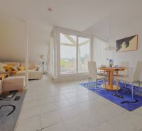 A bright living room of a holiday apartment with a sofa, dining table, and large windows.