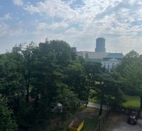 A view of the building among the trees, in the distance Štefánikova Street, behind it offices.