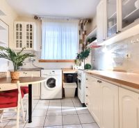 A kitchen in a studio apartment with white cabinets, a washing machine, and a window with white curtains.