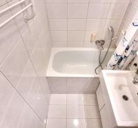A small bathroom in a studio apartment with a bathtub and a sink, tiled with white tiles.