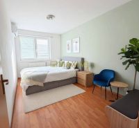 A bright bedroom with a bed, wood-patterned flooring, and furniture in a two-room apartment.