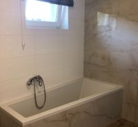 A bathroom in a family house with a bathtub, tiles, and a window. The floor has a wooden decor.