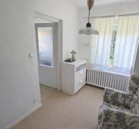 A room in a family house with an armchair, drawers, and a large window with white curtains.