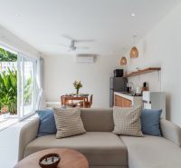 Interior of a villa with a white sofa, kitchen, and view of the pool and greenery.