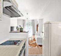 A kitchen with a white fridge, a stove, and a view of the living room in a two-room apartment.