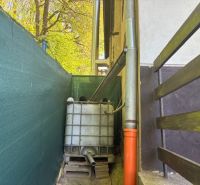 Under the cottage in Ilava, there is a plastic water tank with drainage and green fencing.
