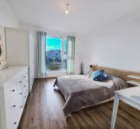 A bright bedroom in a two-room apartment with wood-patterned flooring and a large window.