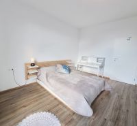 Bedroom in a 2-room apartment with a bed, table, and wooden decor flooring.