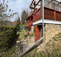 A cottage in Svinica with a stone facade and a wooden terrace, surrounded by greenery.