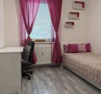 A room in a 4-room apartment with pink curtains, a bed, a table, and a wooden decor floor.