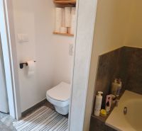 A bathroom in a 4-room apartment with a toilet, shelf, and bathtub, floor with wood decor.