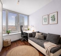 A room with a sofa, a desk, a wooden patterned floor, and large windows.