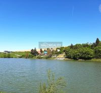 A body of water and houses around the gardens in Hubina, surrounded by greenery and a blue sky.