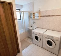 A bathroom in a family house with a washing machine, dryer, and bathtub on light tiles.