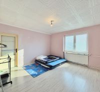 A room in a family house with a mattress on the floor, light pink walls, and a large window.