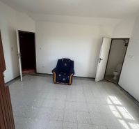 A room with a chair, white walls, tiles, and a door leading to the toilet in the cottage.