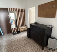 A hallway in a 2-room apartment with a wooden-patterned floor, a mirror, and a black dresser.
