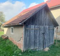 An old wooden garage on a residential plot in Budiná, surrounded by greenery and trees.