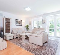 Living room of a family house with a light-colored sofa set, wood-patterned flooring, and large windows.
