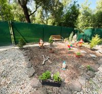 A garden with gnomes and a bench near a cottage in Gabčíkovo, surrounded by stones and a fence.