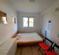 Studio apartment with a bed, white wardrobe, wood-patterned flooring, two folding chairs.