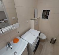A bathroom in a studio apartment with a washing machine, sink, and toilet, light wall tiles.