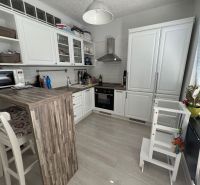 A kitchen in a 2-room apartment with white furniture, a bar counter, and a wooden decor floor.