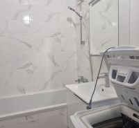 A bathroom in a 2-room apartment with a shower, bathtub, and washing machine, tiled with white tiles.