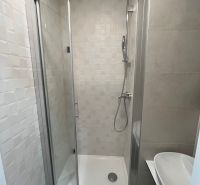 A shower corner and a sink in the bathroom of a studio apartment with simple white tiles.