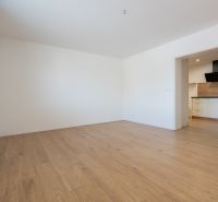 A room in a family house with white walls, a wooden decor floor, and a kitchen.