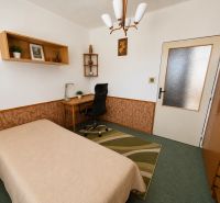 A room in a 3-room apartment with a table, bed, and shelf, green carpet.