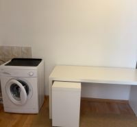 A spacious room in a one-bedroom apartment with a washing machine and a white table, wooden decor flooring.