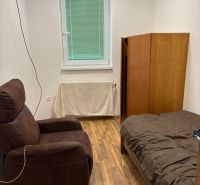 A room with a window, a brown armchair, a bed, and a wardrobe, with a wooden decor floor.