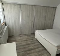 A bedroom in a one-room apartment with a white bed, built-in wardrobe, and wood-patterned flooring.