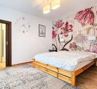 A bedroom with a large bed, bold floral wallpaper, and a wooden-patterned floor.