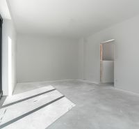 A spacious room with large windows, white walls, and a raw floor. Family house.