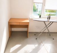 A bright room in a 3-room apartment with a desk, ironing board, and window.