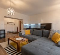 Living room with a kitchen, gray sofa, wooden table, and floor with a wooden design.