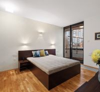 Bedroom in a 2-room apartment with a bed, wood-patterned flooring, and large windows.