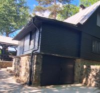 Black cottage in the village of Bukovec with a stone foundation, surrounded by greenery and a shelter.