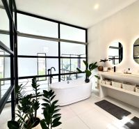 A bathroom in a villa with a bathtub, large windows, round mirrors, and houseplants.