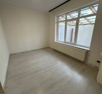 A room in a three-room apartment with a wooden decor floor, white walls, and a large window.
