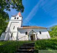 The church in the village of Ozdín with a tower and staircase, surrounded by greenery and a blue sky.