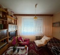 Living room in a two-room apartment with shelves, armchairs, and a window with white curtains.
