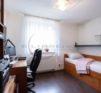 A room in a family house with a bed, a desk, and a wooden-patterned floor.