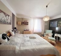 A bedroom in a family house with a bed, paintings, and a chair, floor with wooden decor.