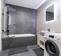 A bathroom in a 3-room apartment with a bathtub, washing machine, and illuminated mirror.