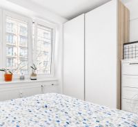A bright bedroom with a large window, white wardrobe, and dresser in a two-room apartment.