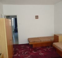 A room in a 2-room apartment with simple furniture and a red carpet.