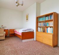 A room in a family house with a chair, a bed, a shelf, and a colorful carpet.